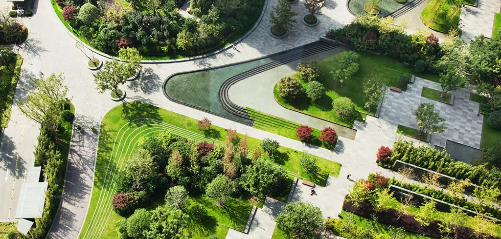 An aerial view of a beautifully landscaped park with manicured lawns, mature trees, and a winding water feature. The park features paved pathways, seating areas, and a variety of plant life